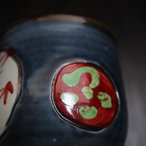 May include: A handcrafted ceramic mug with a dark blue glaze. The mug has two decorative, oval designs. One is white with red and blue accents, the other is red with green spots.