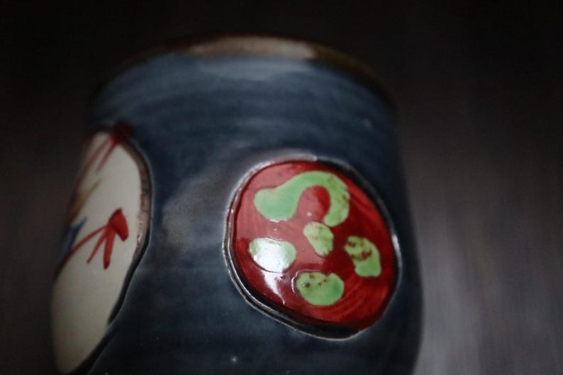 May include: A handcrafted ceramic mug with a dark blue glaze. The mug has two decorative, oval designs. One is white with red and blue accents, the other is red with green spots.