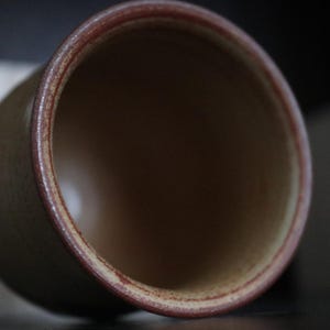 May include: A ceramic cup with a cylindrical form. The cup has a light brown exterior with a darker brown rim. The interior of the cup is a darker brown colour. The cup is tilted slightly, showing the inside.