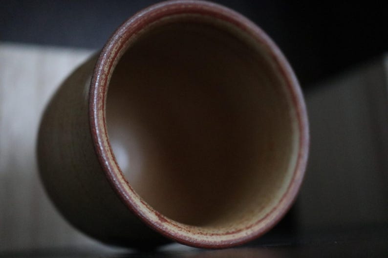 May include: A ceramic cup with a cylindrical form. The cup has a light brown exterior with a darker brown rim. The interior of the cup is a darker brown colour. The cup is tilted slightly, showing the inside.