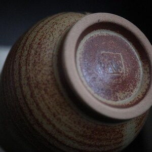 May include: A ceramic vase with a speckled, textured surface in shades of brown and tan. The base is a darker reddish-brown with a lighter rim. The bottom features a stamped mark. Pottery.
