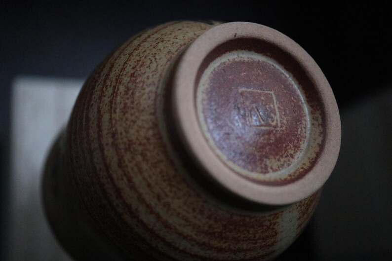 May include: A ceramic vase with a speckled, textured surface in shades of brown and tan. The base is a darker reddish-brown with a lighter rim. The bottom features a stamped mark. Pottery.
