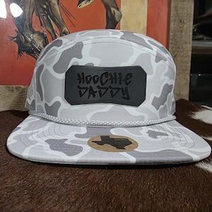 May include: A gray and white camouflage-patterned cap with a flat brim. The cap features a black rectangular patch with the words "HOODIE DADDY" in stylized lettering. A Texas-shaped sticker is on the brim.