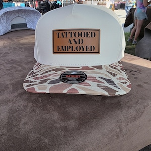 May include: A white baseball cap with a brown leather patch that reads "TATTOOED AND EMPLOYED." The bill of the cap features a camouflage pattern in shades of brown, tan, and cream. The cap is sitting on a brown surface.