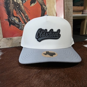 May include: A white baseball cap with a grey brim and a grey, script-style patch on the front that reads Oldschool. A small, tan, Texas-shaped sticker is affixed to the center of the brim, featuring the word Texas printed in black lettering.