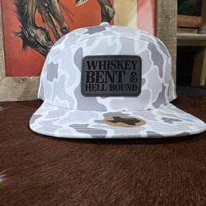 May include: A five-panel baseball cap featuring a light gray and white camouflage pattern. A rectangular black patch on the front displays the text "WHISKEY BENT & HELL BOUND" in a bold font. A small sticker shaped like the state of Texas is on the brim.