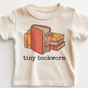 May include: Cream-colored t-shirt featuring a cartoon bookworm wearing glasses, peeking out from behind a red book. The shirt has the words "tiny bookworm" printed in black below the graphic. A stack of books is also visible.
