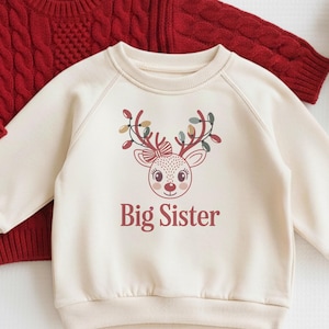 May include: Two children's sweaters and a small stuffed reindeer. One red cable-knit sweater and a cream-colored sweatshirt with a reindeer graphic and the text "Big Sister". The reindeer has brown antlers and a red nose.