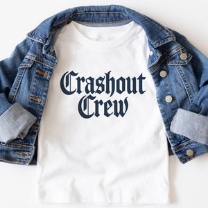 May include: A white t-shirt with the text "Crashout Crew" in a dark blue gothic font. The shirt is paired with a blue denim jacket. The jacket is folded over the shirt.