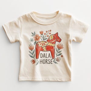 May include: A cream-colored short-sleeve t-shirt featuring a central graphic of a red Dala horse surrounded by muted orange and green floral illustrations. The text DALA HORSE is printed in a dark brown sans-serif font below the horse figure.