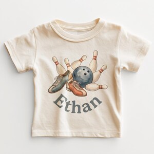 May include: A cream-colored short-sleeve t-shirt featuring a watercolor graphic of a dark gray bowling ball, several beige bowling pins, and a pair of bowling shoes in blue and orange. The name Ethan is printed in dark gray below the graphic.