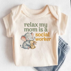 May include: Cream-colored baby onesie with the text "relax my mom is a social worker" in green and yellow. The onesie features a graphic of a gray elephant hugging a tan bunny. Light blue baby pants are visible.