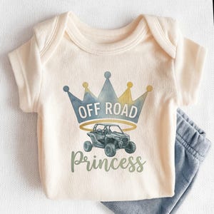 May include: Cream-colored baby onesie with a graphic of an off-road vehicle, a crown, and the words "OFF ROAD PRINCESS" in blue and green. Light blue baby pants are visible on the right side of the onesie.