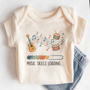 May include: Cream-colored baby onesie with a musical theme. The design includes a guitar, drums, musical notes, and the text "MUSIC SKILLS LOADING...". Light blue baby pants are visible in the image.