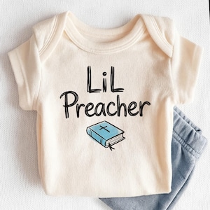 May include: Cream-colored baby onesie with the text "Lil Preacher" in black script. Below the text is a blue book with a cross on the cover. Light blue baby pants are visible at the bottom right.