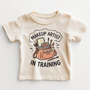May include: A cream-colored short-sleeve t-shirt featuring a graphic illustration of a makeup bag filled with brushes, a mirror, and cosmetics. The text "MAKEUP ARTIST IN TRAINING" is printed in black, bold, rounded lettering above and below the bag.