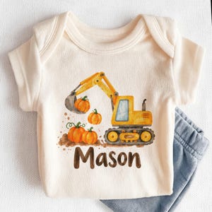 May include: A cream-colored short-sleeve bodysuit featuring a watercolor graphic of a yellow construction excavator scooping orange pumpkins. The name Mason is printed in brown lettering below the excavator. A pair of blue pants is visible.