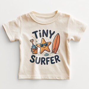 May include: A cream-colored short-sleeve t-shirt featuring a graphic of a smiling starfish wearing sunglasses, a sand bucket with a shovel, and a surfboard. The text "TINY SURFER" is printed in dark blue letters above and below the illustration.