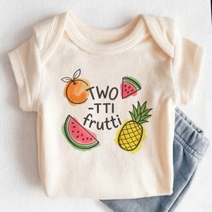May include: Cream-colored baby onesie with the text "TWO -TTI frutti" and illustrations of an orange, watermelon, and pineapple. Light blue pants are visible at the bottom. The onesie is made of soft material.