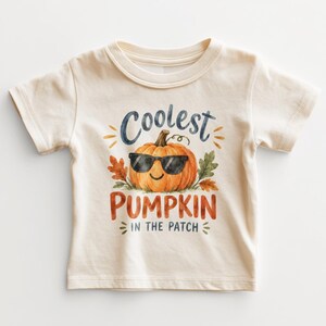 May include: A cream-colored short-sleeve t-shirt featuring a watercolor graphic of an orange pumpkin wearing black sunglasses. The text reads Coolest PUMPKIN IN THE PATCH in blue and orange lettering, surrounded by small orange and green leaves.