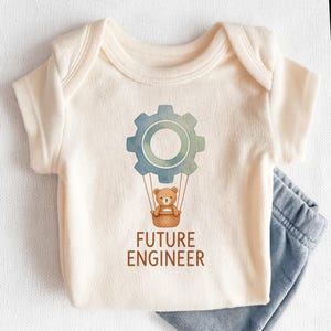 May include: Cream-colored baby onesie with a blue and green gear graphic, a brown teddy bear in a basket, and the text "FUTURE ENGINEER." Light blue baby pants are visible on the right side of the onesie.