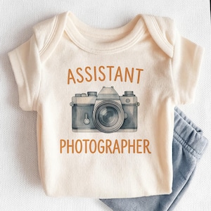 May include: Cream-colored baby onesie with the words "ASSISTANT PHOTOGRAPHER" in brown lettering. A gray and blue camera graphic is centered below the text. Light blue pants are visible at the bottom right.