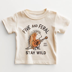 May include: Cream-colored toddler t-shirt with a graphic of a porcupine playing a guitar. The text reads "FIVE AND FERAL" above the porcupine and "STAY WILD" below. The year "EST. 2021" is printed on the right side of the porcupine.