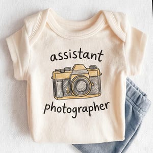May include: Cream-colored baby onesie with a hand-drawn camera graphic in yellow and gray. The text "assistant photographer" is printed below the camera. Light blue baby pants are visible on the right side of the onesie.