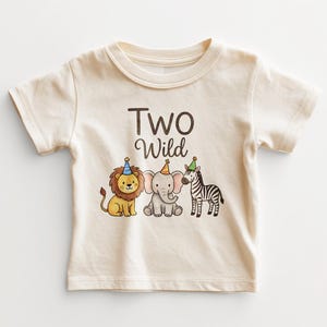 May include: Cream-colored toddler t-shirt with the words "TWO Wild" in brown script. Below the text are illustrations of a lion, elephant, and zebra, each wearing a party hat. The lion is yellow and brown, the elephant is gray, and the zebra is black and white.