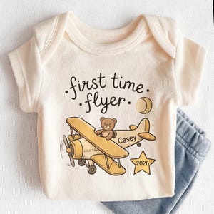 May include: A cream-colored short-sleeve baby bodysuit featuring a graphic of a yellow biplane with a brown teddy bear in the cockpit. The text reads first time flyer, Casey, and 2026 inside a star. A crescent moon is also printed on the fabric.