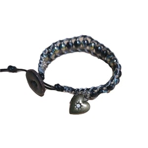 May include: Wrap bracelet with iridescent beads and a small, gold-colored heart charm. The bracelet features a button closure and is made with black and gray cord. The heart charm has a single, clear crystal in the center.