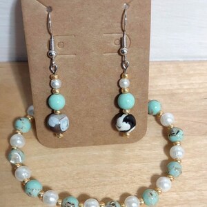 May include: A jewellery set comprising earrings and a bracelet. The earrings have silver hooks, gold accents, and turquoise, black and white, and pearl beads. The bracelet has matching turquoise, pearl, and gold beads.
