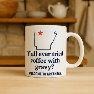 Puede incluir: Taza de cerámica blanca con un contorno negro del estado de Arkansas y una estrella roja. La taza presenta el texto "Y'all ever tried coffee with gravy? WELCOME TO ARKANSAS."