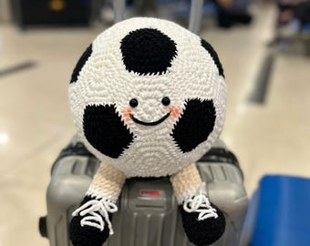 May include: A handmade, crocheted soccer ball plushie with a smiling face, black and white patches, and small legs wearing black shoes with white laces. The plushie is sitting on top of a gray suitcase, suggesting travel or a sports theme.