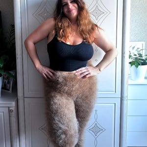 May include: A woman wearing a black tank top and brown, furry leggings. The leggings are the focal point, with a thick, textured appearance. The woman is smiling, with her hands on her hips, posing in front of a white cabinet.