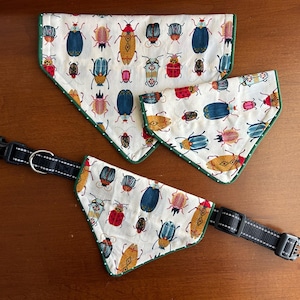 May include: A set of three pet bandanas and a matching collar. The bandanas and collar feature a white background with colorful insect illustrations. The bandanas have a green border. The collar is black with white stitching.