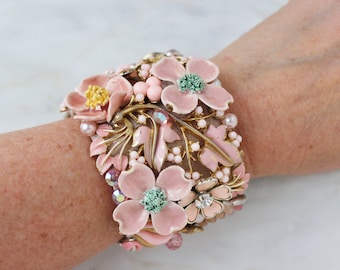 May include: A wide cuff bracelet featuring gold-tone metal leaves and branches adorned with pink enamel flowers, small pink beads, and clear iridescent crystals. The floral design includes textured green centers and light pink petals.