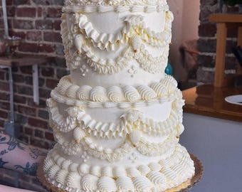 Puede incluir: Un pastel blanco de dos pisos con glaseado de crema de mantequilla elaborado. El pastel presenta volantes decorativos, espirales y detalles dorados. El pastel se asienta sobre una base de cartón dorada, perfecto para una celebración.