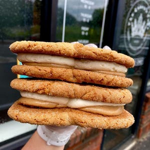 Puede incluir: Una pila de cuatro galletas grandes de color marrón dorado con un relleno de color claro. Las galletas parecen suaves y tienen una superficie ligeramente texturizada. El relleno es una mezcla de blanco y marrón claro, lo que sugiere una crema o glaseado en capas.