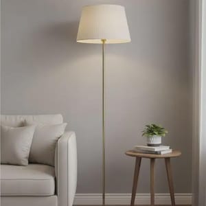 Op de afbeelding: Een goudkleurige vloerlamp met een crèmekleurige lampenkap staat naast een witte bank en een kleine houten bijzettafel. Een kleine potplant staat op de tafel en voegt groen toe aan de kamer. De lamp is ongeveer 160 cm hoog.