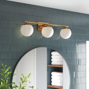 Puede incluir: Una lámpara de tocador dorada de tres luces con pantallas esféricas de vidrio blanco. La lámpara está montada en una pared de azulejos verde azulado sobre un espejo redondo. La decoración del baño incluye una estantería con toallas blancas y una planta verde.