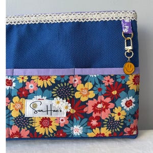 May include: A blue fabric bag with a floral pattern featuring yellow, red, and pink flowers. The bag has a lace trim and a small smiley face charm. The brand name "Sun Hae's" is visible on a label.