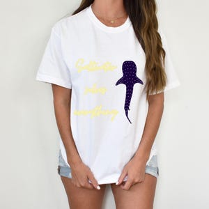 May include: White t-shirt with the text "Saltwater solves everything" in yellow script. A dark blue whale shark graphic with white dots is to the right of the text. The shirt is a casual, unisex style.