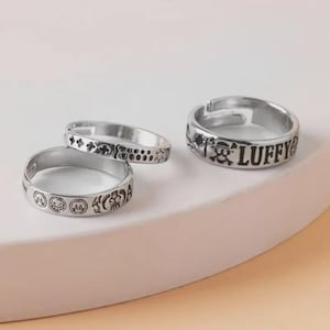 Può includere: Tre anelli d'argento con disegni incisi. Un anello presenta la parola "LUFFY" con un teschio e ossa incrociate. Gli altri due anelli hanno vari simboli e motivi, tra cui facce e disegni floreali. Gli anelli sono esposti su una superficie bianca.