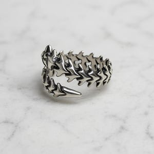 Peut inclure: Une bague argentée en forme d'épine dorsale. La bague est composée de segments détaillés en forme d'os qui s'enroulent autour du doigt. La bague a une finition polie et est posée sur un fond en marbre.