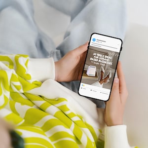 May include: A person holding a smartphone displaying a social media post. The post reads "20 Reels Ideas for Service-Based Businesses." The person wears a white and yellow patterned sweater. The phone screen shows a calendar and white heels.