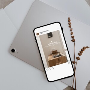 May include: A smartphone displays a social media post with the text "Curated Calm: tools to create a peaceful life." The phone rests on a silver tablet and white paper, with dried flowers. The post shows a desk with a laptop.