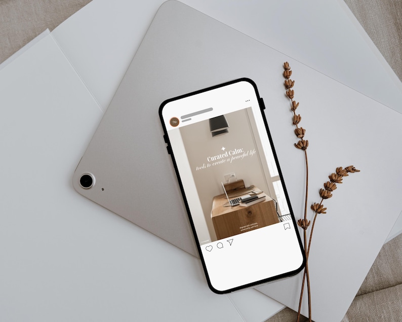May include: A smartphone displays a social media post with the text "Curated Calm: tools to create a peaceful life." The phone rests on a silver tablet and white paper, with dried flowers. The post shows a desk with a laptop.