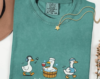 May include: Teal Comfort Colors t-shirt with a graphic of three cartoon ducks engaged in different activities. The text "Just doing my ADLs" is below the ducks. A gold star pendant necklace is draped across the shirt.