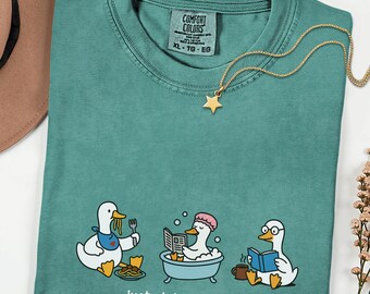 May include: Teal Comfort Colors t-shirt with a graphic of three ducks engaged in daily activities. One eats spaghetti, another bathes, and the third reads. The text "Just doing my ADLs" is below the graphic. A gold star pendant necklace rests on the shirt.